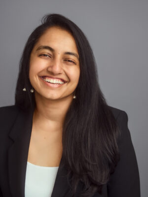 Image description: A brown person with black hair falling past their shoulders looks at the camera, starting to smile. They are wearing a black blazer over a white shirt.