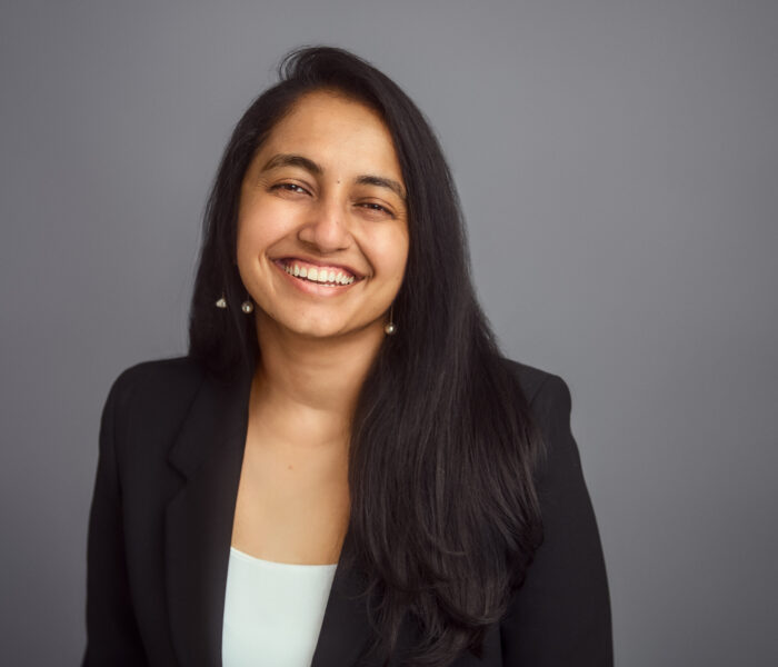Image description: A brown person with black hair falling past their shoulders looks at the camera, starting to smile. They are wearing a black blazer over a white shirt.